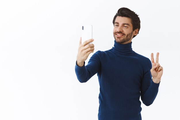 Bel giovane elegante in maglione alla moda collo alto dress-up per la festa di Natale, prendendo selfie per postare online, tenendo in mano lo smartphone, fare gesto di pace e sorridente carino, muro bianco