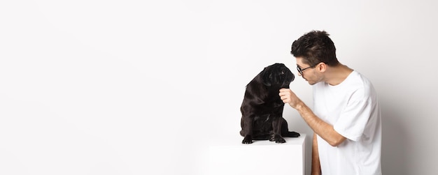 Bel giovane hipster con gli occhiali accarezza il suo cane guardandosi l'un l'altro in piedi su sfondo bianco