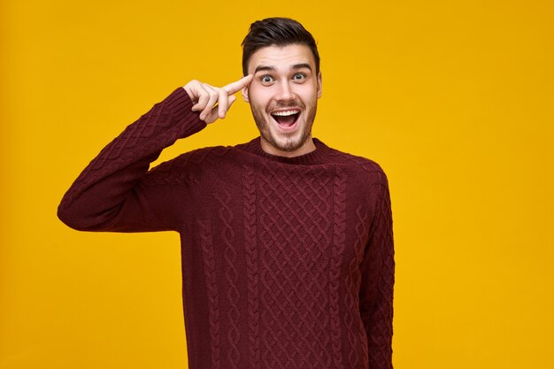 Bel giovane maschio emotivo che esprime Sei un'emozione pazza, tenendo il dito indice vicino alla testa. Ragazzo eccitato in maglione lavorato a maglia rotolando il dito alla tempia, aprendo ampiamente la bocca. Pensa prima di agire