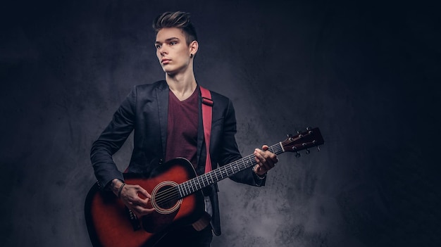 Bel giovane musicista con capelli alla moda in abiti eleganti con una chitarra in mano che suona e posa su uno sfondo scuro.