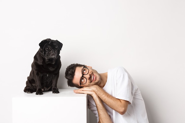 Bel giovane posa la testa vicino a un simpatico carlino nero, sorridendo e guardando lo spazio della copia, sfondo bianco