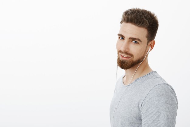 Bel giovane ragazzo caldo maschile e fiducioso con barba e baffi che indossa gli auricolari guardando con bellissimi occhi azzurri audaci e sicuri di sé in posa sul muro bianco