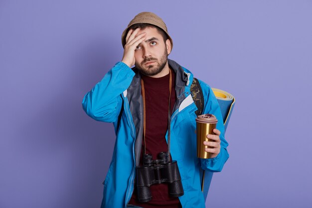 Bel giovane turista maschio europeo che indossa cappello e giacca, avendo il caffè da asporto in tazza termica, sembra stanco, tiene la mano sulla fronte