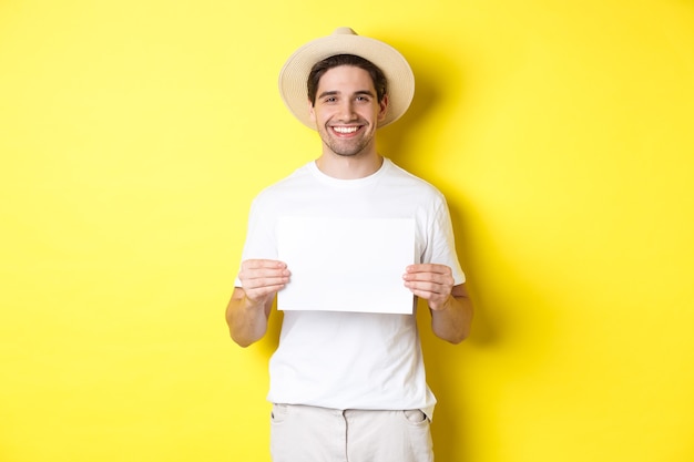 Bel giovane turista maschio in cappello estivo sorridente, con in mano un pezzo di carta bianco per il tuo segno, in piedi su sfondo giallo