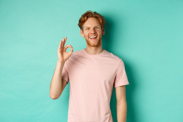 Bel giovane uomo barbuto in t-shirt che mostra il segno Ok, sorride con i denti bianchi e dice sì, d'accordo con te, in piedi su sfondo turchese