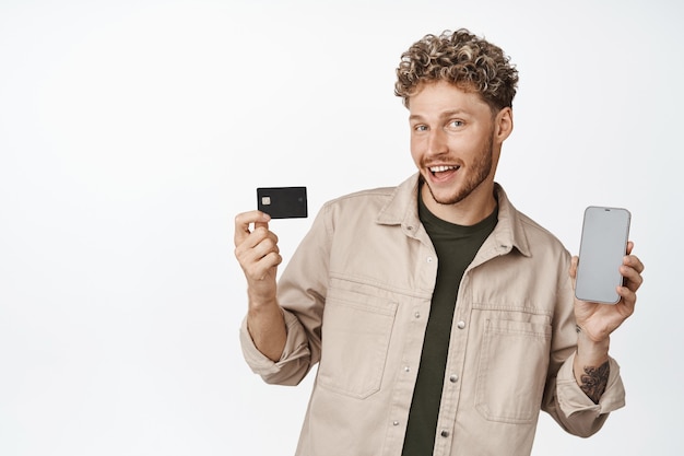 Bel giovane uomo caucasico che mostra la sua carta di credito e lo schermo dello smartphone che mostra un'app che annuncia una nuova applicazione con sfondo bianco
