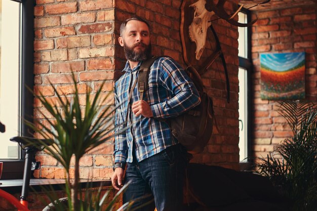 Bel maschio hipster barbuto in una camicia blu in pile e jeans con zaino appoggiato a un muro di mattoni in uno studio con interni soppalcati.