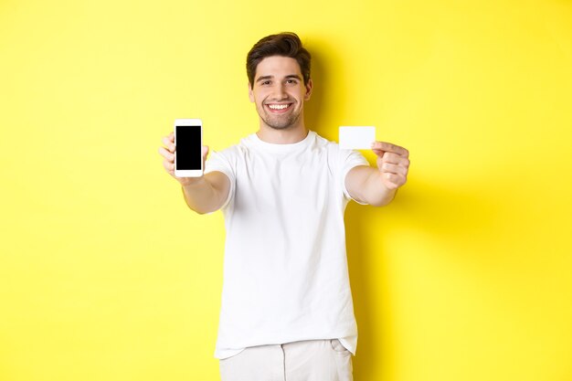 Bel modello maschile caucasico che mostra lo schermo dello smartphone e la carta di credito, il concetto di mobile banking e shopping online, sfondo giallo.