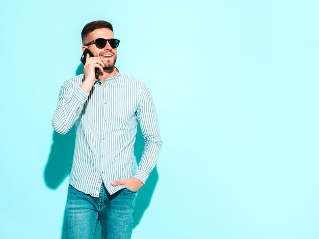Bel modello sorridente Uomo elegante e alla moda che parla allo smartphone Moda uomo hipster in posa vicino al muro blu in studio Telefono con telefono cellulare