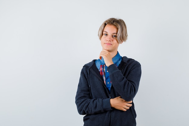 Bel ragazzo adolescente in camicia, felpa con cappuccio che appoggia il mento a portata di mano e sembra allegro, vista frontale.