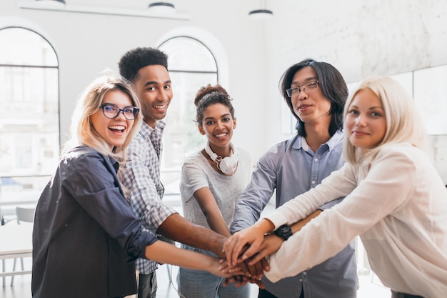 Bel ragazzo africano con l'espressione del viso felice sostenendo i suoi amici prima della conferenza. Ritratto dell'interno del gruppo di lavoro di giovani specialisti internazionali che si preparano all'incontro con il capo.