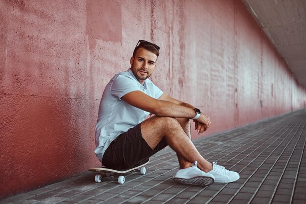 Bel ragazzo alla moda vestito con una camicia bianca e pantaloncini seduto su uno skateboard sotto il ponte, guardando una telecamera.