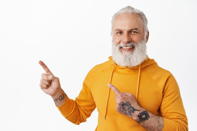 Bel ragazzo anziano con tatuaggi che puntano nell'angolo in alto a sinistra e sorridente dare consigli mostrando pubblicità su uno spazio vuoto in piedi su sfondo bianco