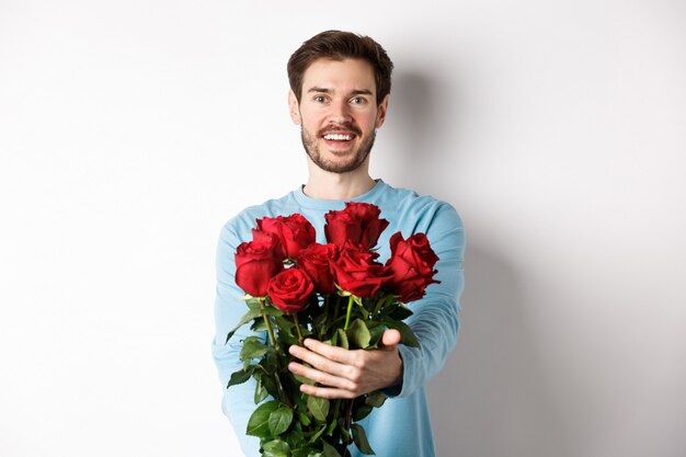 Bel ragazzo barbuto allunga le mani, regala un mazzo di rose e sorride, porta fiori in un appuntamento romantico, festeggia il giorno di San Valentino con l'amante, in piedi su sfondo bianco