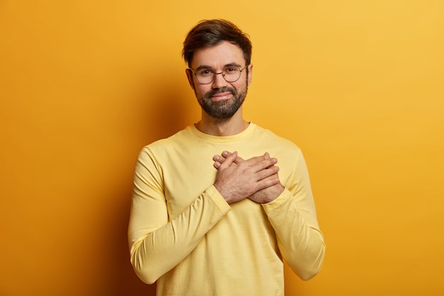 Bel ragazzo barbuto tiene le mani sul cuore, esprime emozioni sincere, apprezza l'aiuto e le parole commoventi, è grato, indossa un maglione giallo casual, posa al coperto. Concetto di linguaggio del corpo