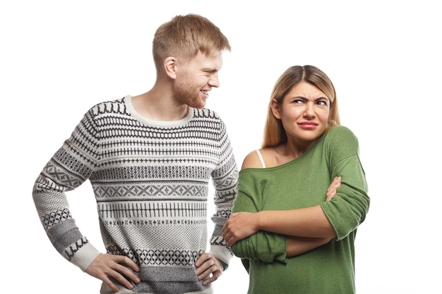 Bel ragazzo barbuto vestito con un maglione sorridente e guardando una donna attraente che sta in una postura chiusa con le braccia incrociate, sentendosi confuso perché non le piace o non capisce la sua stupida battuta