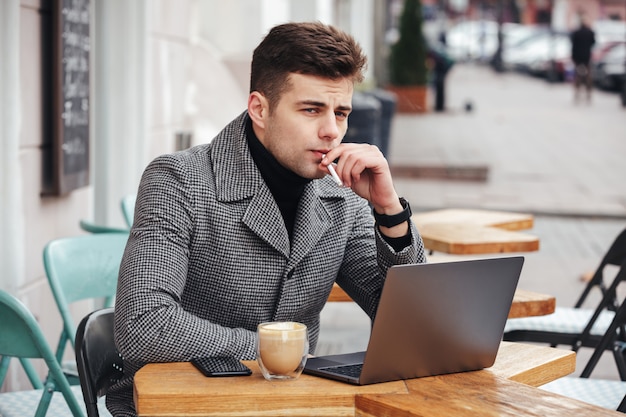 Bel ragazzo caucasico con sguardo meditabondo seduto nella caffetteria fuori, fumare sigarette e bere cappuccino