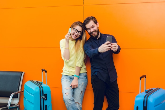 Bel ragazzo con la barba in camicia nera con pantaloni sta facendo selfie-ritratto con una bella ragazza vicino su sfondo arancione tra due valigie. Ha i capelli lunghi, maglione, jeans e parla al telefono