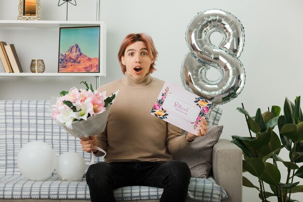 Bel ragazzo confuso il giorno delle donne felici che tiene un bouquet con un biglietto di auguri seduto sul divano in soggiorno