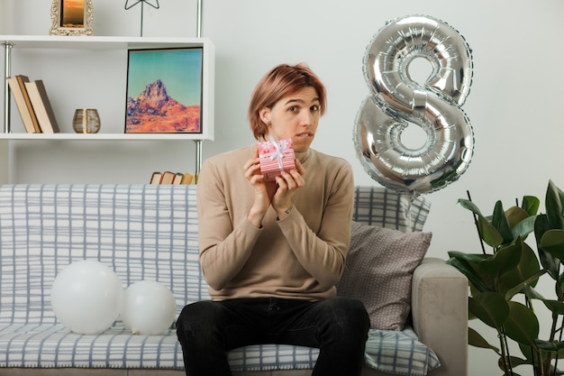 Bel ragazzo confuso nel giorno delle donne felici che tiene il regalo seduto sul divano in soggiorno