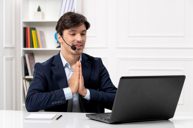 Bel ragazzo del servizio clienti con auricolare e laptop in giacca e cravatta che prega e parla al computer