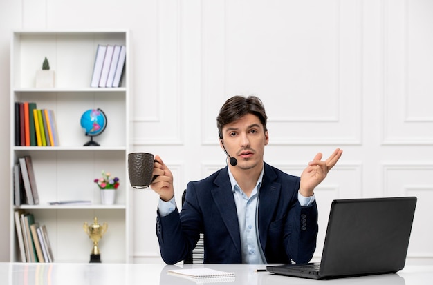 Bel ragazzo del servizio clienti con auricolare e laptop in tuta agitando le mani e tenendo la tazza