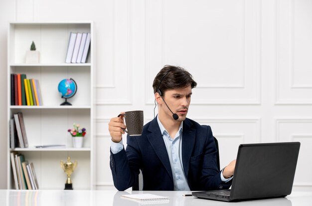 Bel ragazzo del servizio clienti con auricolare e laptop in tuta confuso con la tazza di caffè