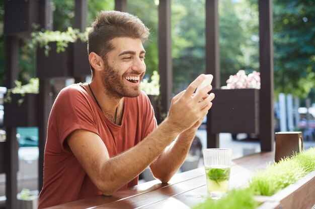 Bel ragazzo felice seduto al bar, bevendo limonata e utilizzando il telefono cellulare, ridendo sul messaggio di testo