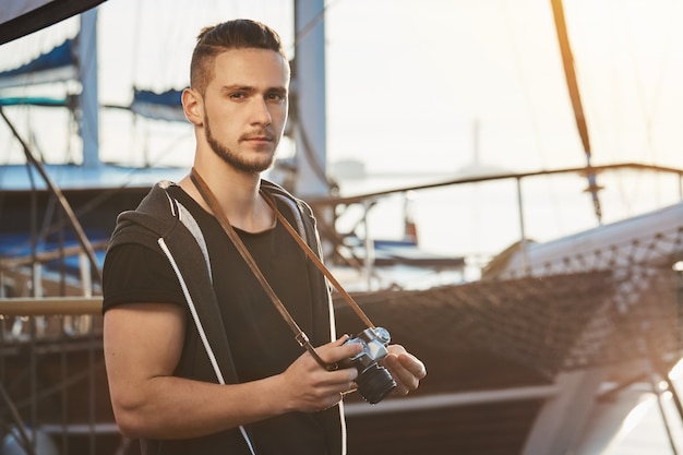 Bel ragazzo fiducioso con taglio di capelli alla moda in piedi vicino al fantastico yacht, tenendo la fotocamera, fissando seriamente e focalizzato durante la sessione fotografica nel porto, facendo foto di paesaggi