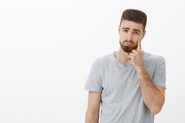 Bel ragazzo giovane toccato con la barba che fa espressione triste e cupa che punta alla palpebra come se mostrasse una lacrima che esprime rammarico o tristezza in piedi scontento piangendo sul muro grigio