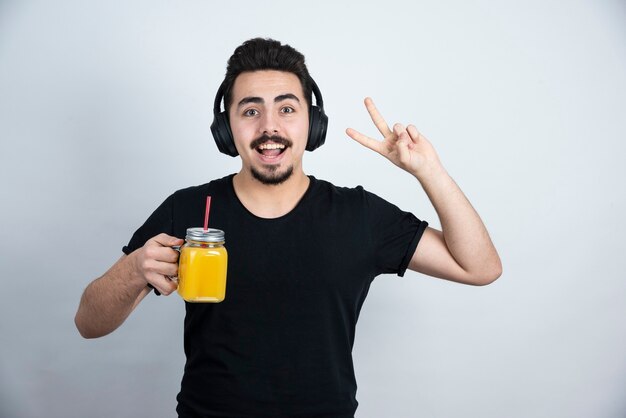 bel ragazzo in cuffia con una tazza di vetro di succo d'arancia che mostra il segno di vittoria.