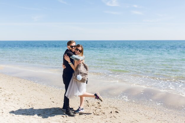 Bel ragazzo in maglietta nera e pantaloni sta abbracciando bella donna con i capelli lunghi vicino al mare