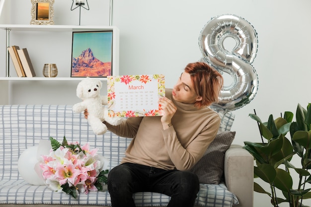 Bel ragazzo in una felice giornata delle donne che tiene e guarda l'orsacchiotto con il calendario seduto sul divano nel soggiorno