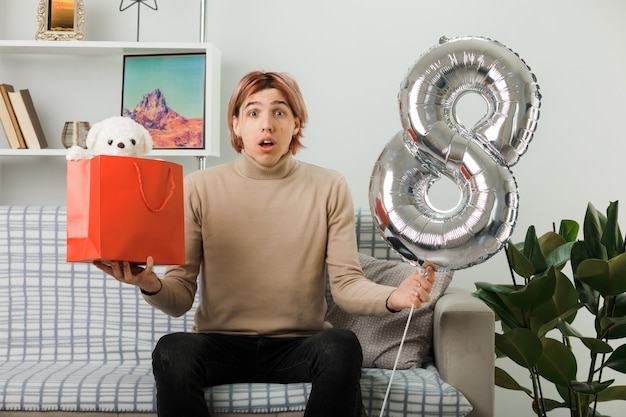 Bel ragazzo in una felice giornata delle donne che tiene il palloncino numero otto con un sacchetto regalo seduto sul divano in soggiorno