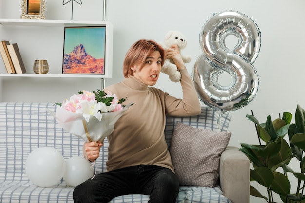 Bel ragazzo in una felice giornata delle donne che tiene in mano un bouquet con un orsacchiotto seduto sul divano in soggiorno