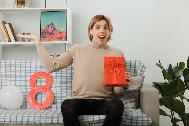 Bel ragazzo in una felice giornata delle donne con un regalo seduto sul divano in soggiorno