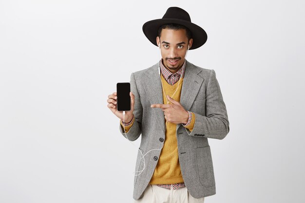 Bel ragazzo nero impertinente in vestito e cappello hipster, ascoltando musica in auricolari, puntando il dito sullo schermo dello smartphone