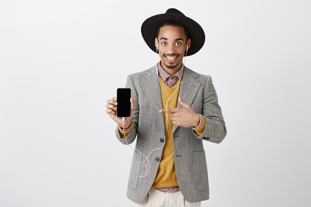 Bel ragazzo nero sorridente in tuta e cappello hipster, ascoltare musica in auricolari, puntare il dito sullo schermo dello smartphone