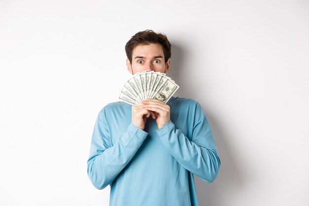 Bel ragazzo sorpreso e stupito che mostra soldi, guardando l'offerta promozionale, facendo shopping con contanti, in piedi su sfondo bianco.