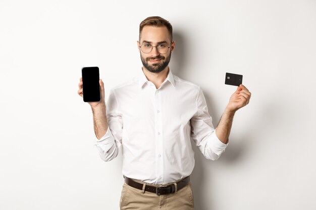 Bel uomo barbuto che mostra il telefono cellulare e la carta di credito, acquisti online, in piedi