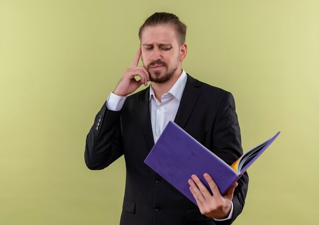 Bel uomo d'affari che indossa tuta azienda cartella blu guardandolo confuso e perplesso in piedi su sfondo verde