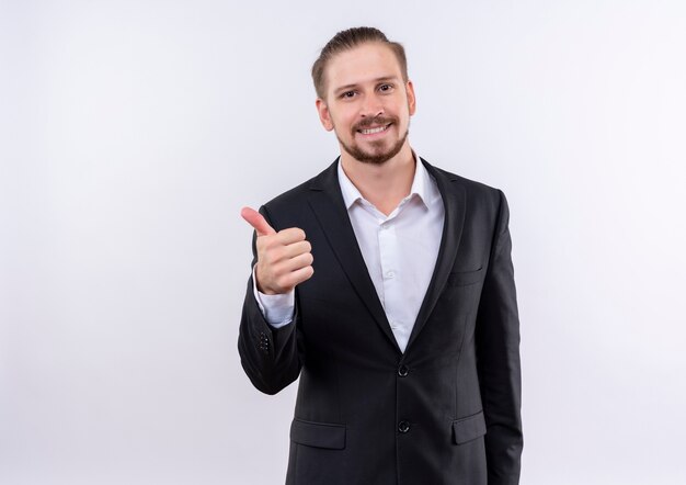 Bel uomo d'affari che indossa tuta guardando la telecamera sorridente che mostra i pollici in su in piedi su sfondo bianco