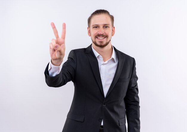 Bel uomo d'affari che indossa tuta guardando la telecamera sorridente che mostra il segno di vittoria in piedi su sfondo bianco