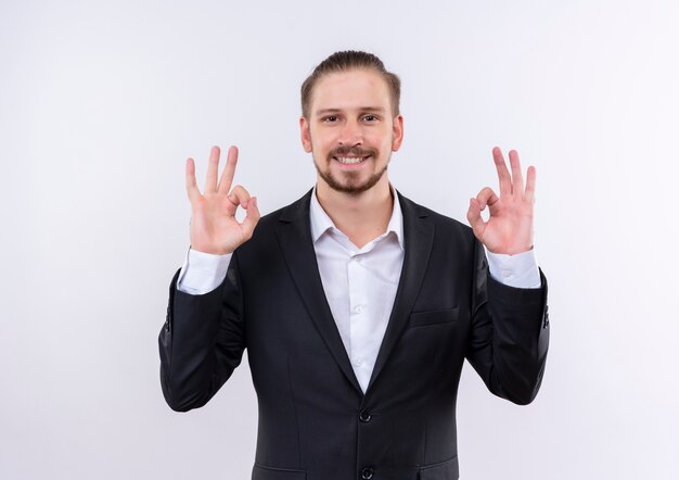 Bel uomo d'affari che indossa tuta guardando la telecamera sorridente che mostra ok cantare con entrambe le mani in piedi su sfondo bianco