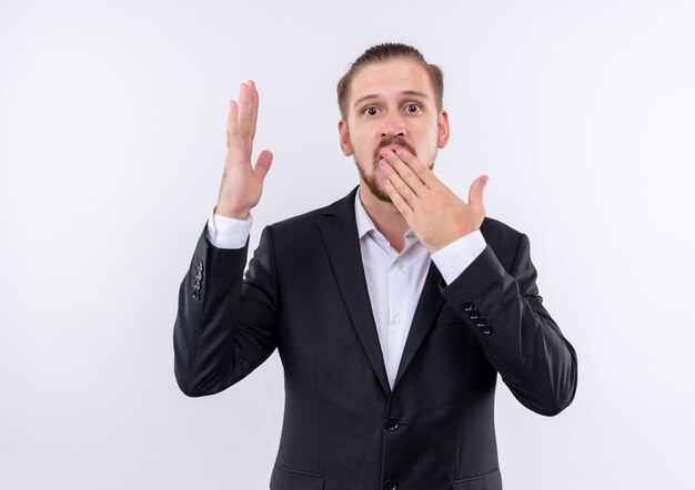 Bel uomo d'affari che indossa tuta guardando sorpreso che copre la bocca con la mano in piedi su sfondo bianco