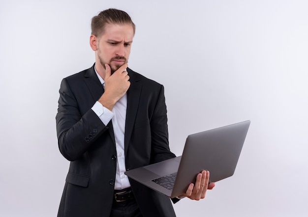 Bel uomo d'affari che indossa tuta tenendo il computer portatile a guardarlo con pensieroso espressione pensando in piedi su sfondo bianco