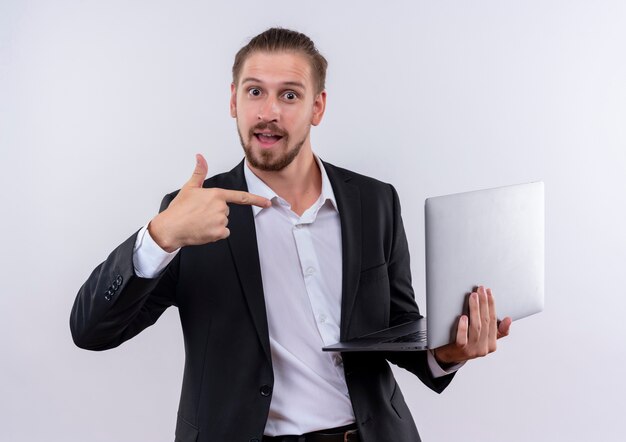Bel uomo d'affari che indossa tuta tenendo il computer portatile che punta con il dito ad esso sorridendo allegramente in piedi su sfondo bianco