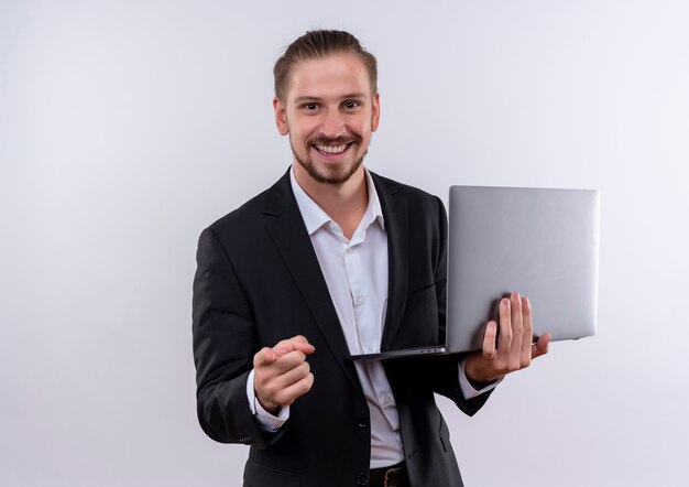 Bel uomo d'affari che indossa tuta tenendo il computer portatile che punta con il dito alla fotocamera sorridente allegramente in piedi su sfondo bianco