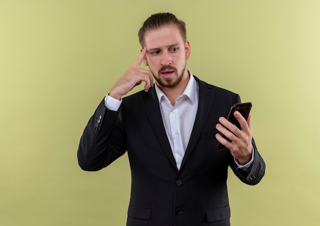 Bel uomo d affari che indossa tuta tenendo lo smartphone guardando lo schermo confuso in piedi su sfondo verde