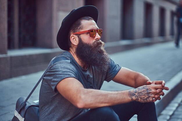 Bel viaggiatore hipster con una barba elegante e un tatuaggio sulle braccia vestito con abiti casual con una borsa, si siede sul marciapiede, riposando dopo un giro in bicicletta.